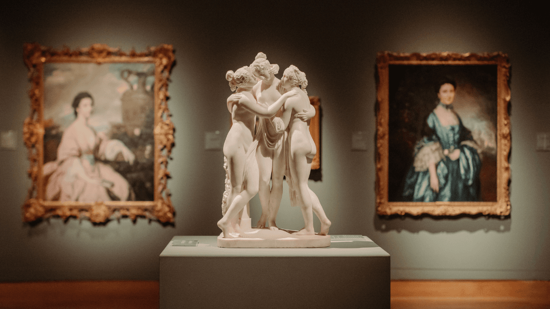 a small white sculpture of three people embraced and two paintings in the background
