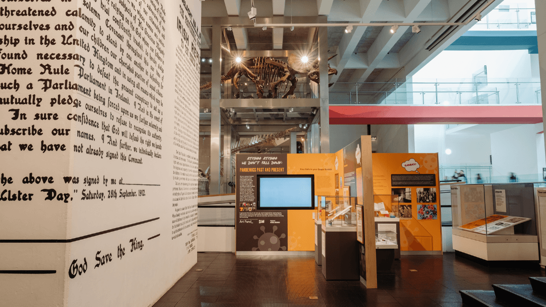 Black and white writing of a large wallpaper poster on the left, yellow and red block walls with TV screens inside an exhibition and brown-coloured dinosaur skeleton in the distance
