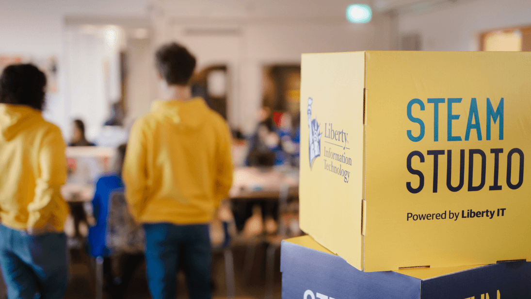 Yellow and navy cubes stacked on top of each other, the yellow cube branded 'STEAM Studio'. Two adults wearing yellow hoodies are visible in the background, with their backs to the camera, where school kids are also sat at a table.