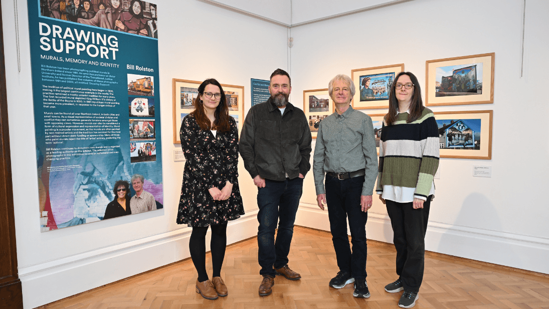 Four people standing inside the Belfast Room of Ulster Museum, two men and two women, at the reveal of a new murals photography exhibition. Framed photographs are on white walls behind them as well as a turquoise wall graphic that says 'Drawing Support'.