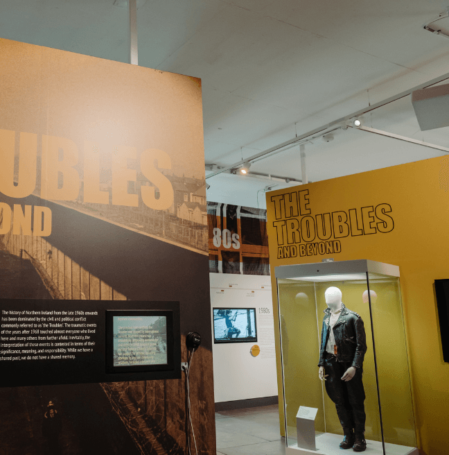 Yellow and black graphics of the Troubles and Beyond exhibition in Ulster Museum. One panel has a built in TV screen and against another wall is a glass cabinet with a jacket on display.